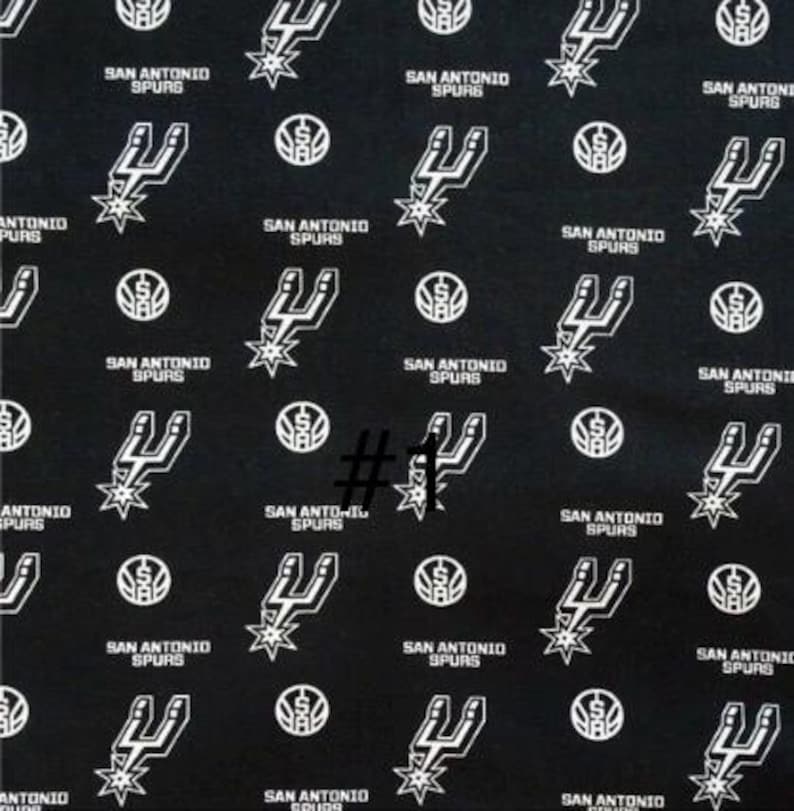 K&ouml;nnte beinhalten: Schwarzer Stoff mit einem sich wiederholenden Muster des Logos und Textes der San Antonio Spurs. Das Design umfasst das Teamlogo, einen stilisierten Sporn mit einem Stern und die Worte "SAN ANTONIO SPURS" in Wei&szlig;.