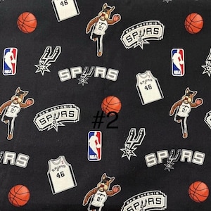 K&ouml;nnte beinhalten: Schwarzer Stoff mit einem sich wiederholenden Muster aus Logos des Basketballteams San Antonio Spurs, Trikots, Basketb&auml;llen und dem Teammaskottchen. Das Trikot hat die Nummer 46. Das NBA-Logo ist ebenfalls vorhanden.