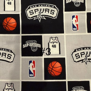 K&ouml;nnte beinhalten: Stoff mit sich wiederholendem Muster mit San Antonio Spurs-Logos, Basketb&auml;llen und Trikots mit der Nummer 46. Das Design enth&auml;lt das NBA-Logo in Rot, Wei&szlig; und Blau, vor schwarzen und grauen Quadraten.