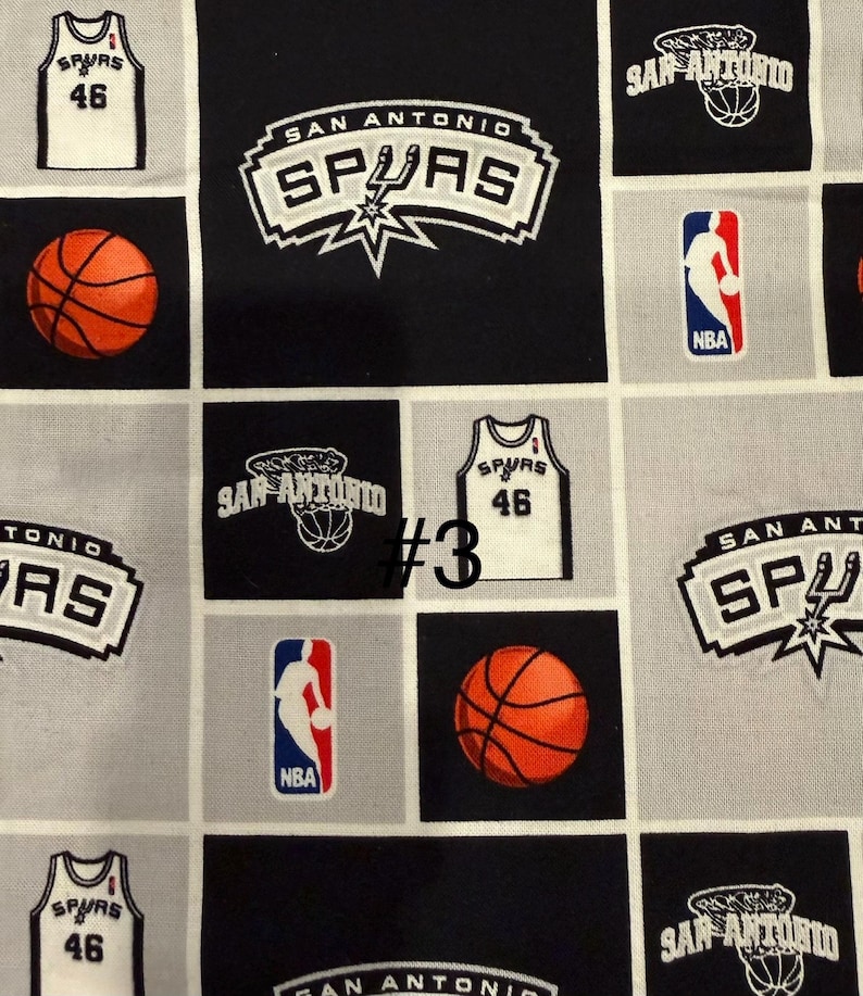 K&ouml;nnte beinhalten: Stoff mit sich wiederholendem Muster mit San Antonio Spurs-Logos, Basketb&auml;llen und Trikots mit der Nummer 46. Das Design enth&auml;lt das NBA-Logo in Rot, Wei&szlig; und Blau, vor schwarzen und grauen Quadraten.
