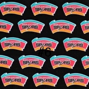 K&ouml;nnte beinhalten: Ein Muster aus San Antonio Spurs Logo-Patches auf schwarzem Hintergrund. Jeder Patch zeigt ein rotes und blaues Design mit dem Text "SAN ANTONIO SPURS". Die Nummer "#6" ist auf einem der Patches sichtbar.