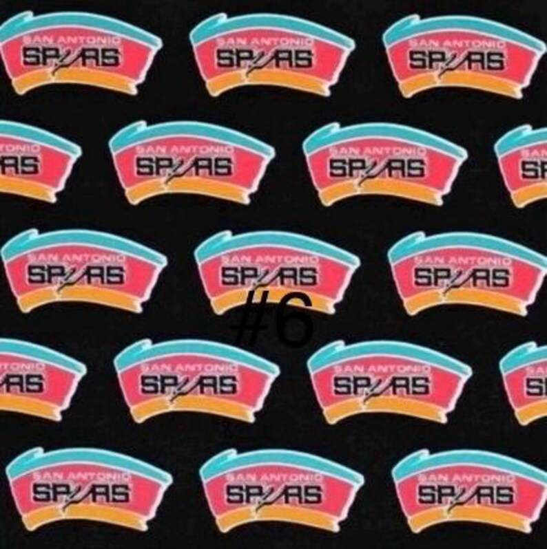 K&ouml;nnte beinhalten: Ein Muster aus San Antonio Spurs Logo-Patches auf schwarzem Hintergrund. Jeder Patch zeigt ein rotes und blaues Design mit dem Text "SAN ANTONIO SPURS". Die Nummer "#6" ist auf einem der Patches sichtbar.