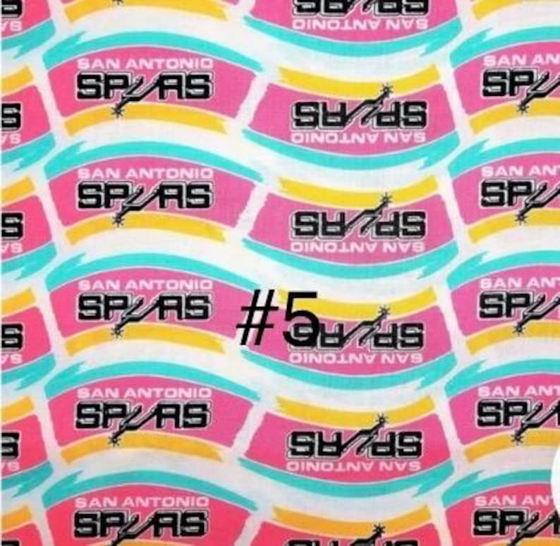 K&ouml;nnte beinhalten: Stoff mit sich wiederholendem Muster des Logos der San Antonio Spurs. Das Design zeigt den Namen und das Logo des Teams in Schwarz, Wei&szlig;, Rosa, Gelb und T&uuml;rkis. Die Nummer "5" ist ebenfalls sichtbar.