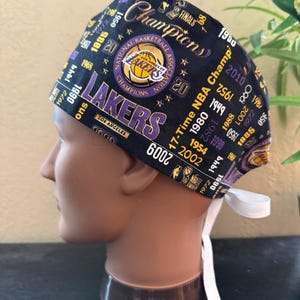May include: Black scrub cap featuring the Los Angeles Lakers logo and championship years. The cap includes the text "Champions" and "Lakers" in gold and purple. A white tie is attached to the back of the cap.