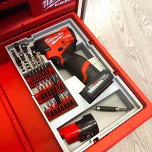 Puede incluir: Una caja de herramientas roja Milwaukee Packout con un inserto gris que contiene un taladro FUEL rojo y negro, brocas y accesorios. La caja de herramientas está abierta, mostrando las herramientas bien organizadas. El logotipo de Milwaukee es visible.