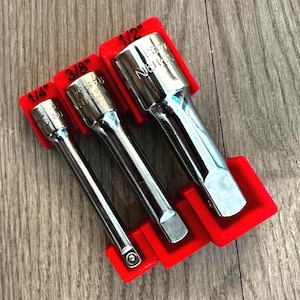 Può includere: Tre chiavi a bussola cromate in supporti rossi. Le chiavi sono etichettate 1/4", 3/8" e 1/2". Gli utensili sono disposti su una superficie di legno.