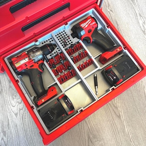 Op de afbeelding: Een rode gereedschapskist met een grijs interieur, georganiseerd met elektrisch gereedschap en accessoires. De gereedschappen omvatten een boor, slagschroevendraaier en diverse bits. De gereedschappen zijn rood en zwart, met de merknaam "Milwaukee" zichtbaar.
