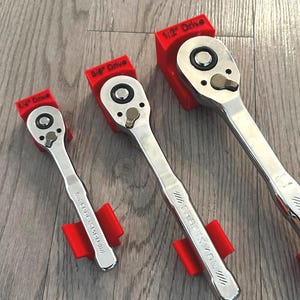 Könnte beinhalten: Drei Craftsman-Ratschen mit roten Haltern. Die Ratschen sind mit 1/4", 3/8" und 1/2" Drive beschriftet. Die Werkzeuge sind silberfarben mit schwarzen Akzenten und werden auf einer hellgrauen Holzoberfläche präsentiert.