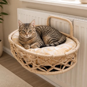 Puede incluir: Un gato atigrado descansa dentro de una cama para radiador de cuerda trenzada beige. La cama presenta una estructura de celosía y un cojín suave de color crema con un sutil patrón circular, enganchada de forma segura a un radiador blanco.