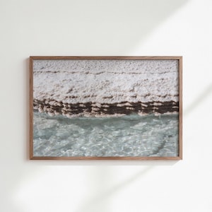 Puede incluir: Impresión enmarcada que presenta un paisaje costero. La imagen muestra un acantilado texturizado y claro sobre agua turquesa transparente. El marco de madera añade un toque natural a la obra de arte, ideal para la decoración del hogar.