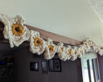 Op de afbeelding: Een gehaakte slinger van crème en gele bloemen hangt boven een deuropening. De handgemaakte bloemenversiering heeft bloemachtige bloemen met gele centra en crèmekleurige bloemblaadjes. De slinger is gemaakt van dik garen.
