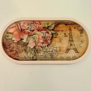 May include: An oval-shaped tray with a light pink border. The interior features a decorative collage with pink peony flowers, a silhouette of the Eiffel Tower, and the text "retro postale" printed in a dark brown script on a tan background.