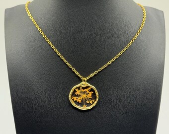 May include: A gold-colored necklace with a circular pendant. The pendant features a dried orange flower encased in clear resin. The necklace is displayed on a black mannequin.