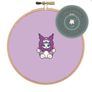 Könnte beinhalten: Ein Kreuzstich-Stickrahmen mit einer pixeligen Katze, die eine lila Kapuze trägt. Die Katze ist weiß, grau und rosa. Der Hintergrund ist helllila. Ein runder Aufkleber mit japanischem Text und einer Katzengrafik ist angebracht.