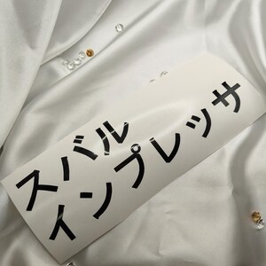 Peut inclure: Un autocollant blanc avec des caractères japonais noirs est présenté sur une surface blanche et soyeuse. L'autocollant indique "Simple Press" en japonais. De petits bijoux décoratifs clairs et dorés sont dispersés autour de l'autocollant.