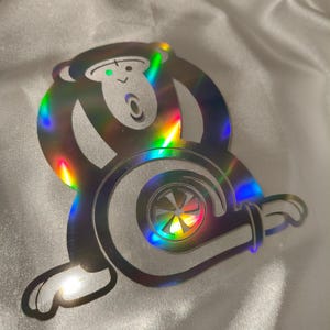 May include: A holographic sticker featuring a cartoon monkey design integrated with a turbocharger. The sticker has a reflective, rainbow-like sheen, with the monkey's face and the turbocharger's details clearly visible. The sticker is placed on a white, textured surface.