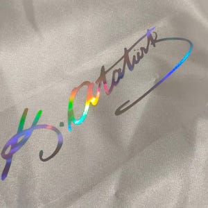 Op de afbeelding: Een holografische sticker met een regenboogkleurige handtekening. De handtekening, leesbaar als "K.Atatürk", staat op een transparante, rechthoekige achtergrond. De sticker ligt op een wit, getextureerd oppervlak.