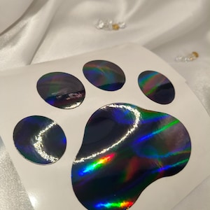 May include: A sheet of stickers featuring a holographic paw print design. The paw print is composed of five circular pads and a larger, rounded pad, all displaying a spectrum of colors including green, blue, and red. The stickers have a glossy finish.