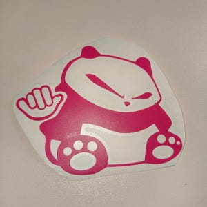 Puede incluir: Una pegatina de vinilo rosa de un oso panda de dibujos animados con una pata levantada, haciendo un signo shaka. El panda está sentado con las piernas cruzadas y tiene un contorno blanco.