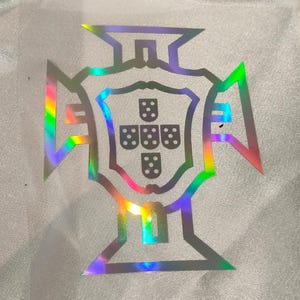 Może przedstawiać: Holograficzny herb Portugalii, przedstawiający tarczę z pięcioma mniejszymi tarczami, z których każda ma pięć kropek. Emblemat umieszczony jest na białym, teksturowanym tle z tęczowym połyskiem.