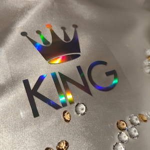 Puede incluir: Tela plateada con una corona holográfica y la palabra «KING» en una fuente a juego. Varios diamantes de imitación transparentes y dorados están dispersos alrededor del texto, añadiendo brillo. Ideal para ropa o accesorios.