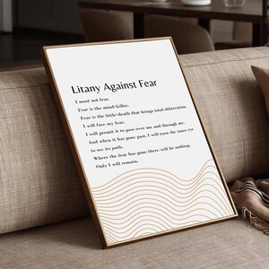 May include: A framed print with the text "Litany Against Fear" in black font. The print has a gold frame and a white background with a wavy, beige design. The print is displayed on a beige sofa with a brown pillow and a patterned blanket.