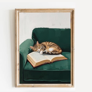 Op de afbeelding: Een ingelijste print van een kat die slaapt op een open boek op een groene fauteuil. De kat heeft bruine en bruine strepen. Het boek heeft vergeelde pagina's en een rode rug.