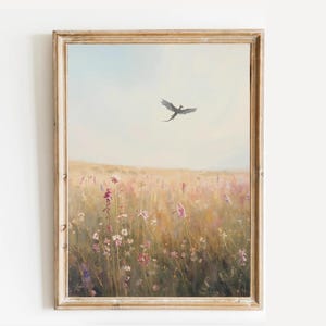 Op de afbeelding: Een ingelijst landschapsschilderij toont een veld met wilde bloemen in roze en gele tinten onder een lichtblauwe lucht. Een donker silhouet van een vliegend wezen is zichtbaar. Het schilderij zit in een goudkleurige houten lijst.
