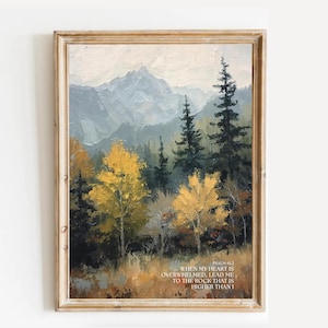May include: A framed landscape painting featuring a mountain range in the background and a forest in the foreground. The trees display shades of green and yellow. The painting includes text at the bottom: "WHEN MY HEART IS OVERWHELMED, LEAD ME TO THE ROCK THAT IS HIGHER THAN I."