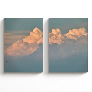 May include: A diptych wall art set featuring two rectangular canvases displaying a continuous sky scene. The images show voluminous, soft-edged clouds illuminated by a warm, golden light against a cool blue sky, with small dark birds in flight.