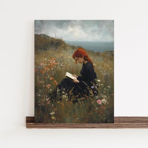 May include: A framed art print depicts a woman with red hair reading a book while seated in a field of wildflowers. The woman wears a black dress, and the background shows a cloudy sky and a body of water. The artwork is mounted on a wooden frame.
