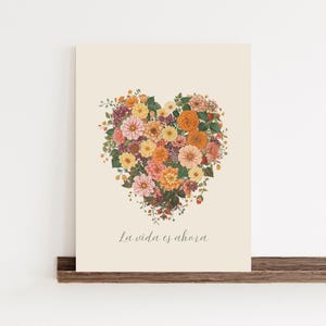 Könnte beinhalten: Ein floraler Kunstdruck mit einer herzförmigen Anordnung bunter Blumen in Rosa-, Orange-, Gelb- und Lilatönen. Der Druck hat den Text "La vida es ahora" unten, vor einem cremefarbenen Hintergrund.