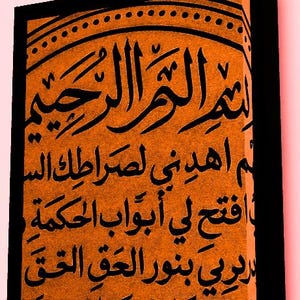 Puede incluir: Impresión en lienzo naranja con caligrafía árabe negra. El texto está centrado y llena el lienzo, con un borde negro. La impresión tiene una apariencia texturizada, y la caligrafía está en escritura tradicional.