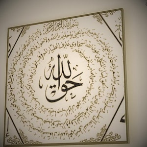 Op de afbeelding: Vierkant ingelijst kunstwerk met Arabische kalligrafie in zwart op een crèmekleurige achtergrond. Het centrale ontwerp bevat een prominent woord, omringd door cirkelvormige tekst. De lijst is in een gedempte goudkleur, wat een vleugje elegantie toevoegt.