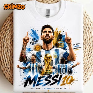 Può includere: Una t-shirt bianca con una grafica di Lionel Messi con la maglia dell'Argentina, il trofeo della Coppa del Mondo e schizzi di vernice blu e oro. Il testo include "LA PULGA", "UN SUEÑO. UN PAÍS. UNA LEYENDA.", "MESSI 10" e "ARGENTINA".