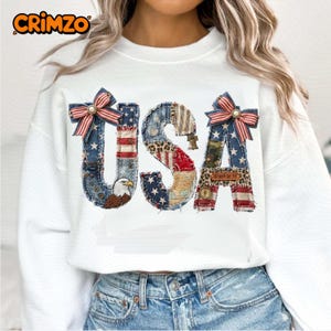 May include: White sweatshirt featuring a patchwork "USA" design. The letters incorporate stars, stripes, and animal print. Each letter is decorated with a red, white, and blue bow. An eagle graphic is included in the design.
