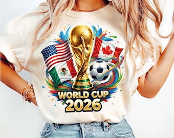 Może przedstawiać: Kremowa koszulka z złotym trofeum Mistrzostw Świata, piłką nożną i flagami USA, Kanady i Meksyku. Koszulka ma złoty napis "WORLD CUP 2026". To element odzieży codziennej.