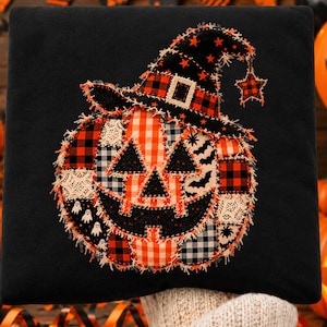 Puede incluir: Un cojín cuadrado negro con una aplicación central de calabaza hecha de varios parches de tela en cuadros naranjas, negros y blancos, encaje y patrones de fantasmas. La calabaza lleva un sombrero de bruja a juego con una estrella colgante.