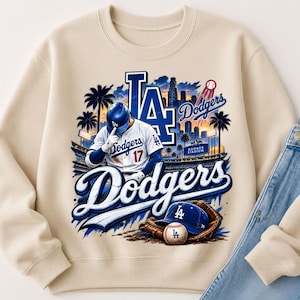 Peut inclure: Un sweat-shirt beige à col rond avec une impression graphique représentant un joueur de baseball en uniforme blanc portant le numéro 17, un logo LA bleu, le texte Dodgers, une silhouette de stade, des palmiers, une casquette bleue et une balle.