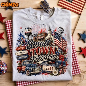 Puede incluir: Una camiseta gris jaspeado con un estampado gráfico de una escena de pueblo pequeño con un camión antiguo, una torre de agua, una iglesia, una bandera estadounidense y un buzón. El texto dice "Small Town Freedom", "Faith", "Family", "Freedom" y "12345".