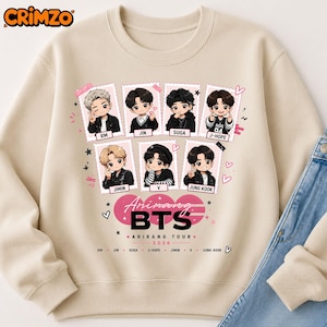 Peut inclure: Un sweat-shirt beige à col rond avec une impression graphique de sept personnages de style chibi dans des cadres photo individuels. Sous les cadres, le texte indique Arirang BTS ARIRANG TOUR 2026, avec les noms des membres et des petits cœurs.