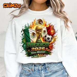 Puede incluir: Una sudadera blanca con un estampado gráfico del trofeo de la Copa Mundial de la FIFA, un balón de fútbol y la bandera de México. El diseño incluye llamas, mazorcas de maíz y el texto "MEXICO 2026 Viva México! CHAMPIONS OF THE WORLD".