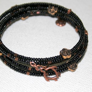 Könnte beinhalten: Ein Wickelarmband mit schwarzen und kupferfarbenen Perlen. Das Armband hat einen kupferfarbenen Verschluss und dekorative Blumenanhänger. Das Armband ist so konzipiert, dass es sich mehrfach um das Handgelenk wickelt.