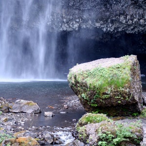 Può includere: Un grande masso ricoperto di muschio si trova di fronte a una cascata. L'acqua scende a cascata da una scogliera rocciosa. L'acqua è limpida e blu, e le rocce sono grigie e marroni.