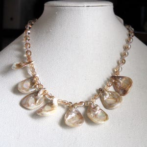 Puede incluir: Un collar hecho de cuentas marrones con colgantes de concha iridiscente. El collar se muestra en un maniquí blanco.