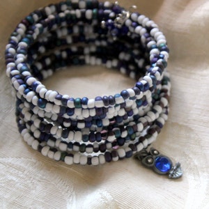 May include: A multi-strand bracelet made of white and blue beads with a small owl charm. The owl charm has a blue gemstone eye.