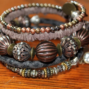 May include: A stack of mixed metal bracelets with various textures and embellishments. The bracelets feature beads in copper, bronze, and silver tones, along with textured fabric and decorative metalwork. The overall aesthetic is bohemian.