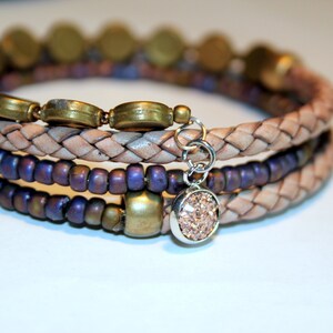 May include: A multi-strand bracelet featuring a braided leather band, purple beads, gold-toned metal beads, and a charm with a sparkly pink stone.
