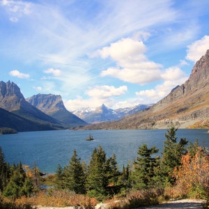 Può includere: Una vista panoramica di un lago circondato da montagne e alberi. Il lago è calmo e blu, con una piccola isola al centro. Le montagne sono ricoperte di alberi e neve, e il cielo è azzurro chiaro con nuvole bianche.