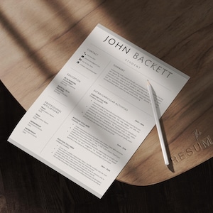 May include: A resume titled "JOHN BACKETT STUDENT" with a white pencil on a wooden surface. The resume includes sections for education, summary, and extracurricular activities. The wooden surface has the word "RESUMADE" engraved on it.